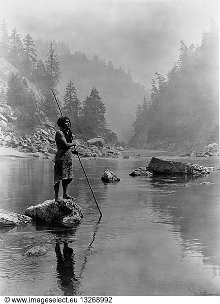 Hupa man with spear  standing on rock midstream  in background  fog partially obscures trees on mountainsides  1923. Photograph by Edward Curtis (1868-1952).