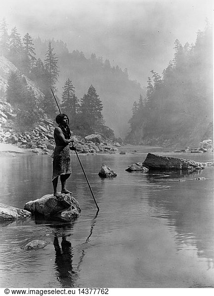Hupa man with spear  standing on rock midstream  in background  fog partially obscures trees on mountainsides  1923. Photograph by Edward Curtis (1868-1952).