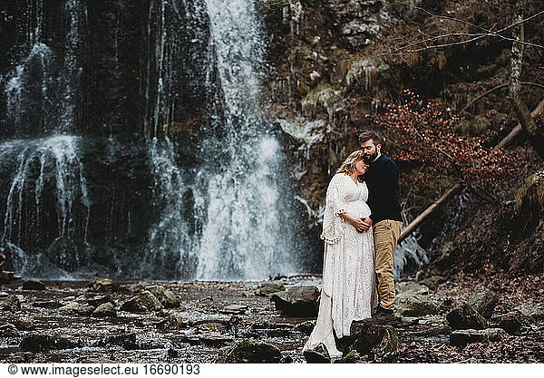Husband and expecting wife outside in winter by a waterfall in winter