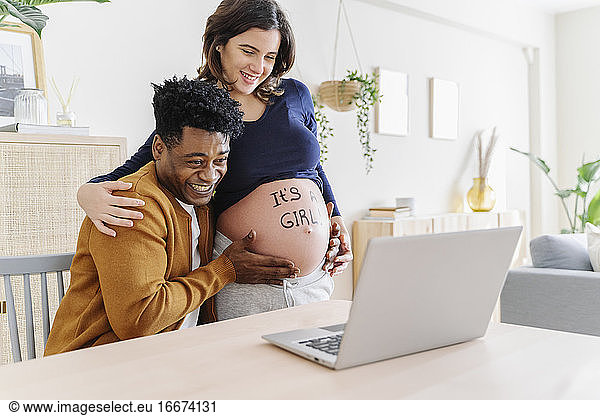 Husband and pregnant wife celebrating ultrasound results in front of a laptop  with a phrase written on her belly that it is a girl. Parenthood concept