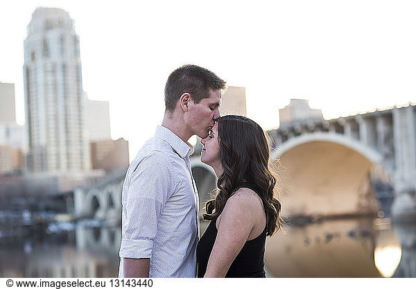 Husband kissing on pregnant wife's forehead while standing by river against clear sky in city