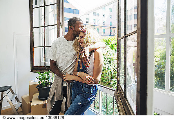 Husband kissing wife on forehead while standing by window in new house