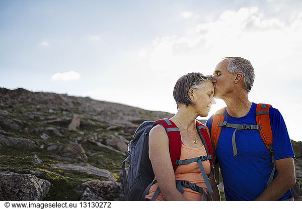 Husband kissing wife on forehead while standing on mountain against sky