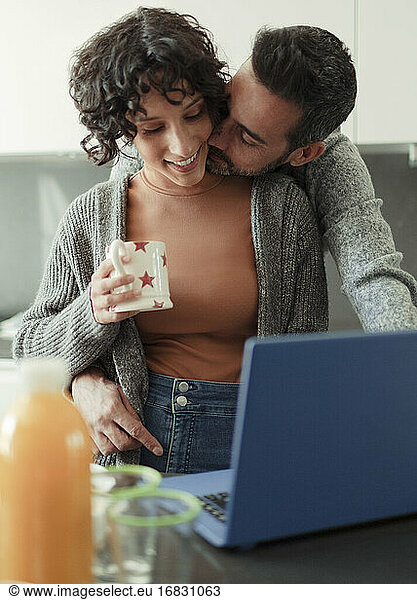 Husband kissing wife working from home at laptop in kitchen
