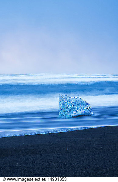 Ice chunk on the shore of the ocean with waves crashing against the shoreline; Iceland