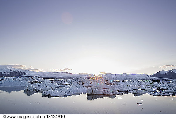 Icebergs by snowcapped landscape during sunrise against clear sky