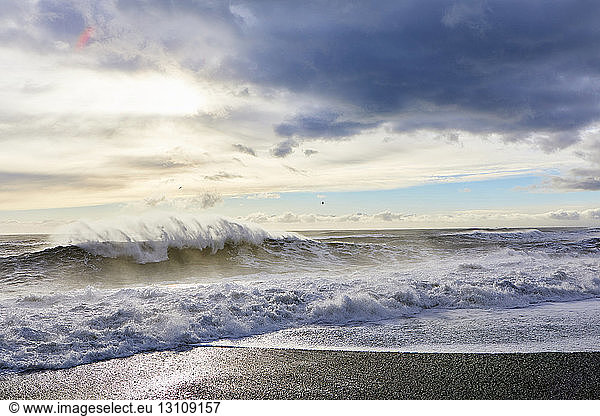 Idyllic view of waves splashing on shore against cloudy sky
