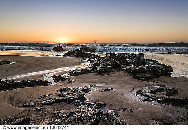 Idyllischer Blick auf das Meer mit Felsen am Strand vor dramatischem Himmel bei Sonnenuntergang