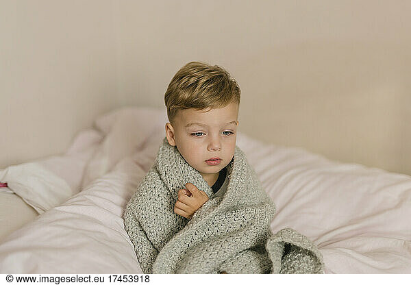 Ill and tired boy wrapped in grey blanket sitting on bed