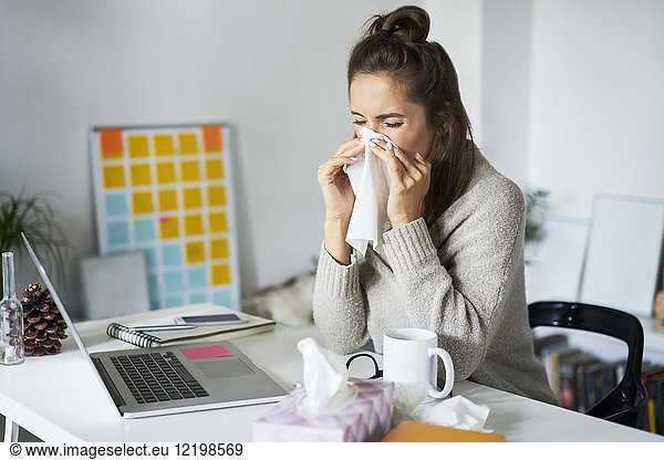 Ill woman at home at desk blowing her nose
