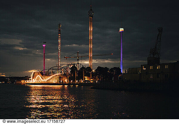 Illuminated amusement park by sea against sky at night