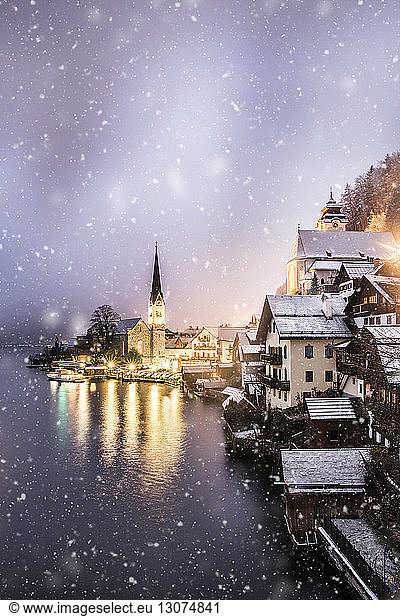 Illuminated buildings by lake against sky during snowfall at sunset