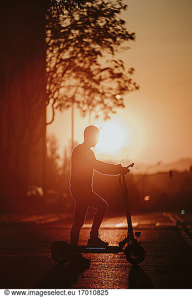 In silhouette of man standing with electric push scooter on road during sunset in city