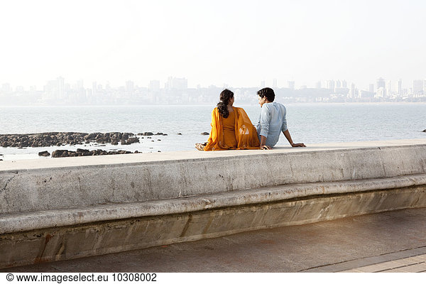India  Mumbai  Young couple sitting at beach promenade  looking at ocean