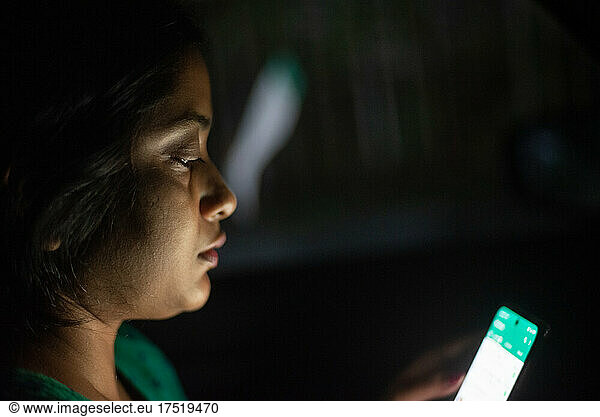 Indian woman looking at her cellphone sitting inside a car in darkness