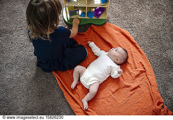 Infant on orange blanket watching sister play with toys