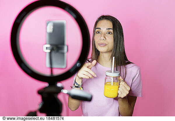 Influencer with orange juice vlogging on mobile phone over pink background