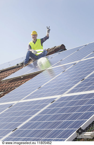 Ingenieur macht eine Friedensgeste  während er eine Pause von der Installation von Solarzellen auf dem Hausdach macht