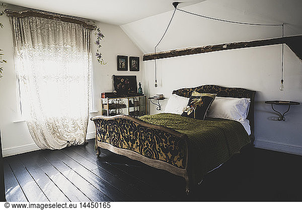 Interior view of attic bedroom with dark wooden floor and white walls  antique French double bed with green throw and lace net curtain in window.