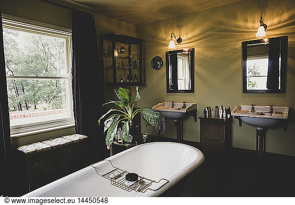 Interior view of bathroom with black mirrors over two Victorian wash stands  sash window and roll top bath with brass bath caddy.