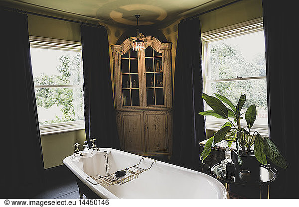Interior view of bathroom with wooden corner cabinet between sash windows  roll top bath with brass bath caddy.