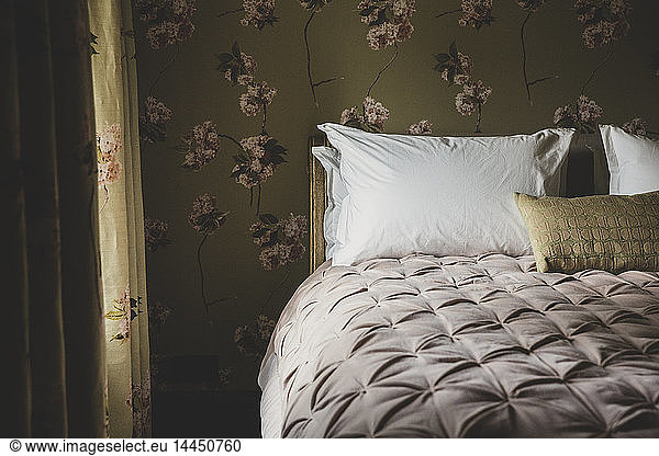 Interior view of bedroom with curtains and wallpaper with floral pattern  pale pink quilt and white pillows on double bed.