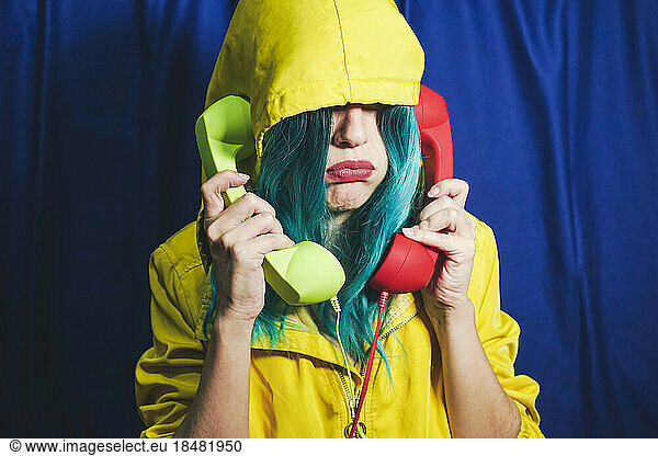 Introvert woman in hooded shirt talking on green and red telephones in front of blue backdrop