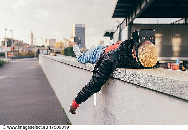 Italy  Woman with VR goggles lying on wall in city