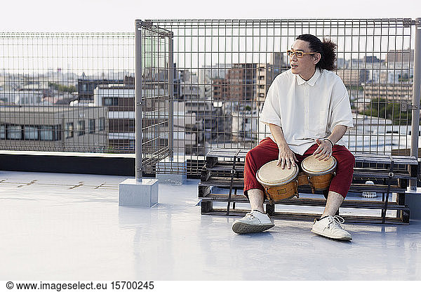 Japanese man sitting on a rooftop in an urban setting  playing drums.