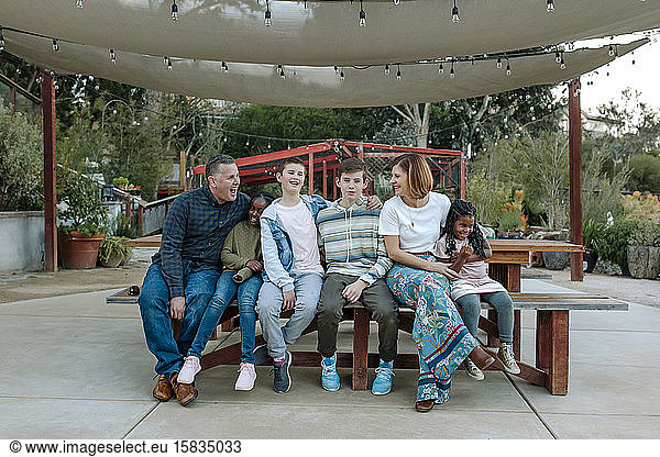 Joyful multiracial family laughing on bench under sunshade at nursery