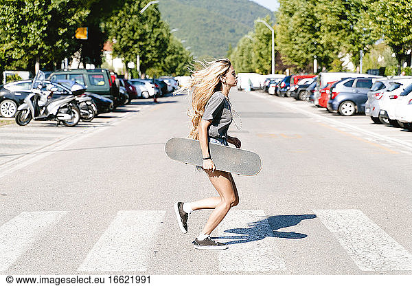 Junge blonde Frau hält Skateboard  während sie über einen Zebrastreifen in der Stadt läuft