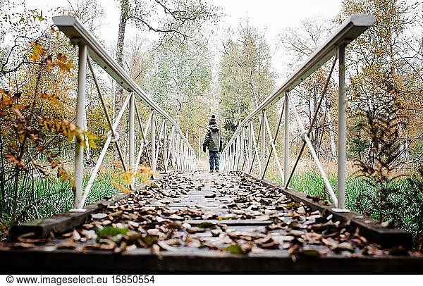 Junge  der im Herbst im Wald eine Brücke mit Herbstlaub entlanggeht