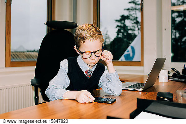Junge  der in einem Büro arbeitet und bei der Arbeit auf das Telefon seines Vaters schaut