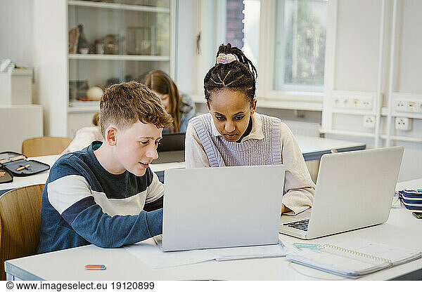 Junge  der sich einen Laptop mit einer Freundin teilt  die am Schreibtisch im Klassenzimmer sitzt