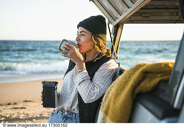 Junge Frau  die ein Getränk in einem Becher genießt  während sie allein im Auto am Strand campiert