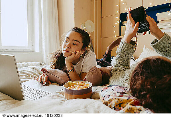 Junge Frau  die einen Laptop benutzt  während ihr Freund auf dem Bett eine Textnachricht über sein Smartphone sendet