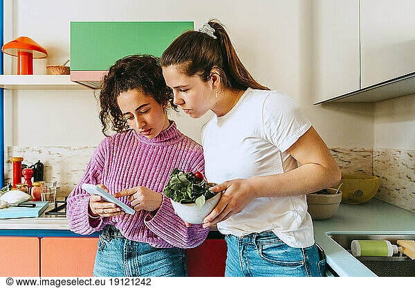 Junge Frau  die ihr Handy mit einer Freundin teilt  während sie sich zu Hause auf die Küchentheke stützt