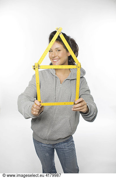 Junge Frau hält ein aus einem gelben Zollstock geformtes Haus in den Händen und lacht in die Kamera