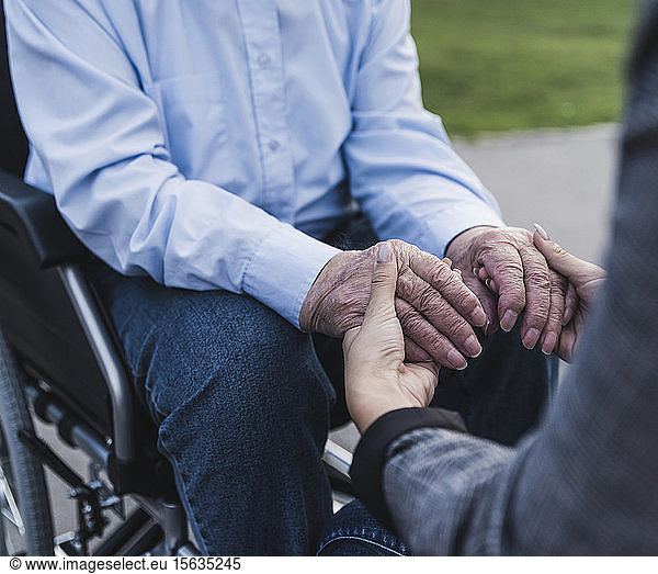 Junge Frau hält Hände eines älteren Mannes  der im Rollstuhl sitzt  Teilansicht