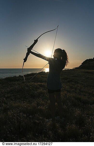 Junge Frau hält Pfeil und Bogen bei Sonnenuntergang