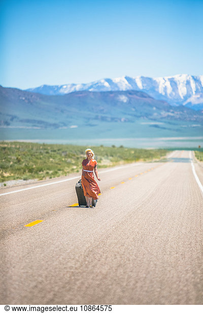 Junge Frau im Stil der 1950er Jahre  die allein auf dem Highway 50 geht und einen Rollkoffer zieht  Nevada  USA