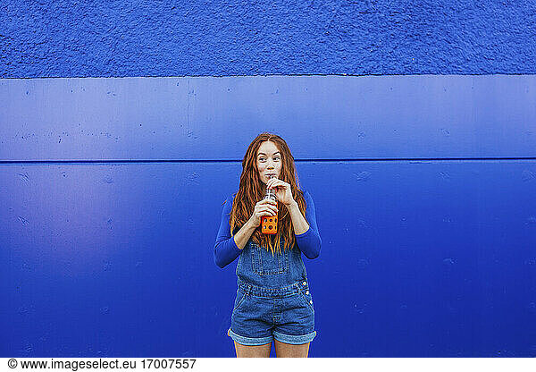 Junge Frau in blauer Freizeitkleidung trinkt Saft  während sie an einer blauen Wand steht