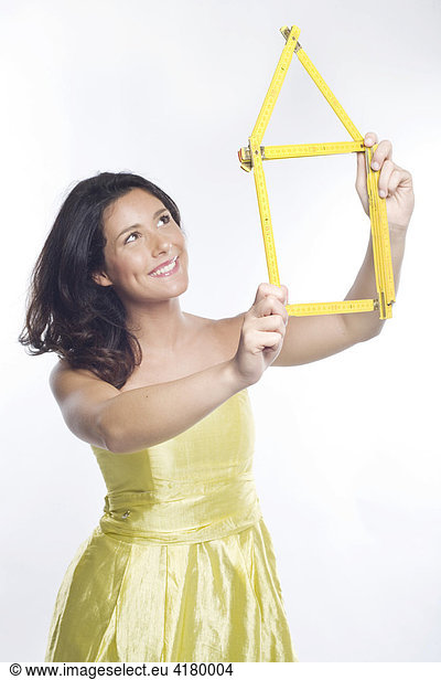 Junge Frau in gelbem Kleid hält lachend ein aus einem gelben Zollstock geformtes Haus in den Händen