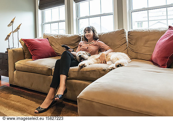 Junge Frau in Geschäftskleidung sitzt mit Hund auf der Couch und sieht fern.