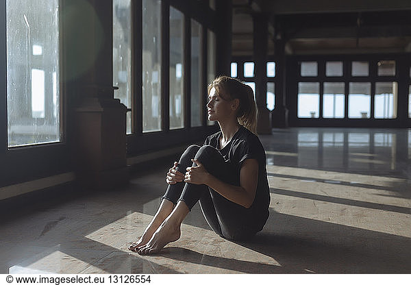 Junge Frau in voller Länge mit geschlossenen Augen  die die Knie hält  während sie auf dem Fliesenboden sitzt