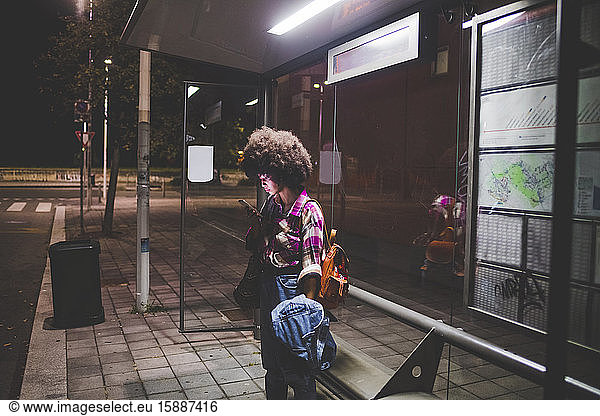 Junge Frau mit Afro-Frisur benutzt Smartphone an Bushaltestelle in der Stadt