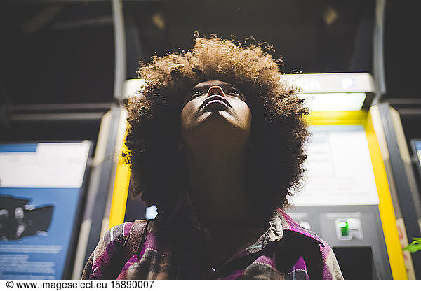 Junge Frau mit Afro-Frisur nachts am Fahrkartenautomaten aufblickend