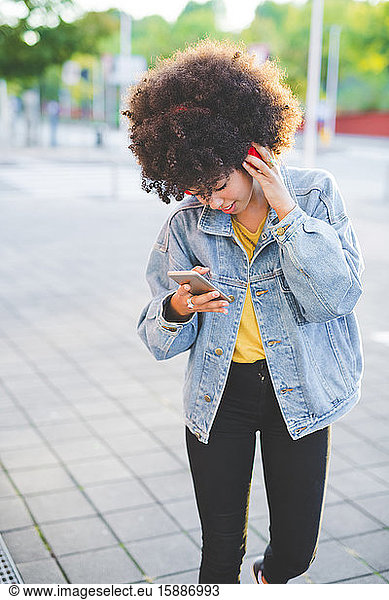 Junge Frau mit Afrofrisur benutzt Smartphone und Kopfhörer in der Stadt