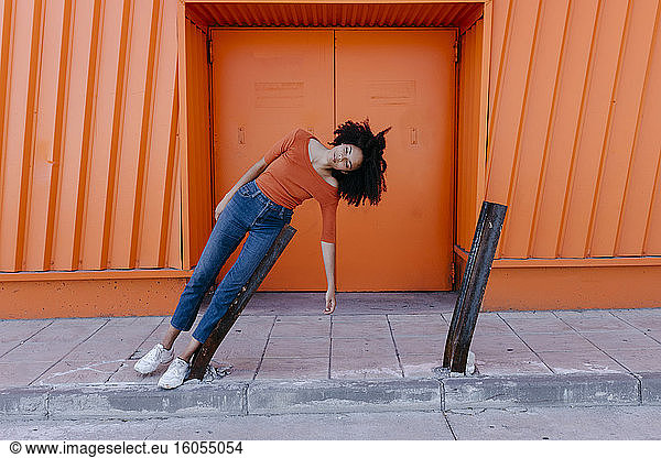 Junge Frau mit Afrofrisur lehnt an beschädigtem Metall gegen orangefarbene Tür
