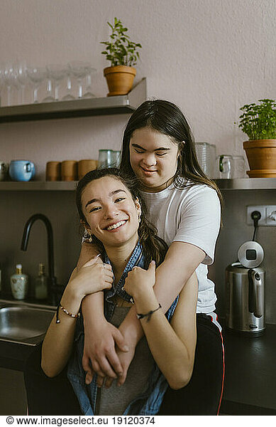 Junge Frau mit Down-Syndrom umarmt Schwester von hinten in der Küche zu Hause
